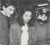 A photograph from the Super Mario-A-Thon event showing Justine Bateman (star of Family Ties) playing Super Mario Bros. while being cheered on by her brother, Jason Bateman (star of Valerie) and a person in a Mario costume cheer for her.