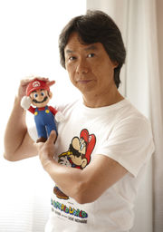 Shigeru Miyamoto holding a Mario figurine. He is also wearing a Super Mario World t-shirt.