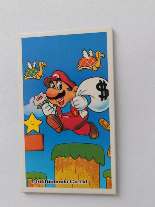 A Nintendo trading card depicts Mario jumping over some grassy platforms while holding a sack over his shoulder. The sack is labeled with a dollar sign. Some golden coins appear to be spilling out of the sack. In the background is a Starman atop an empty block, and two Koopa Paratroopas, one red and one green.