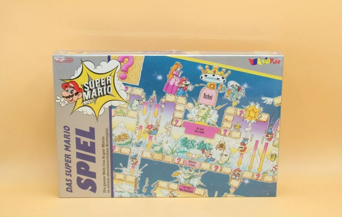 A sealed board game box is displayed. On the left is a large grey vertical stripe with "Das Super Mario Spiel" written vertically across it, the Nintendo logo, and artwork of Mario's head and hand flashing a peace sign next to a Super Mario Bros. logo. The rest of the box depicts a complex scene in Super Mario Land's Easton Kingdom, with various Marios falling through layers of blocks or confronting various enemies and bosses from Super Mario Land. Neat the top of the image, Princess Peach stands behind Mario as he shoots a fireball at Tatanga, with the fight taking place under a royal crown.