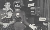 A photograph from the Super Mario-A-Thon event showing winner Wil Wheaton and his younger brother, Jeremy, with a person in a Mario costume. The grand prize and sweepstakes prizes are shown right next to them.