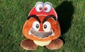 Official photograph of a Goomba plush toy wearing Cappy and mustache cut-outs from a printable